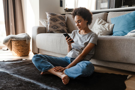 Young beautiful smiling curly african woman holding and using her phone sitting on carpet on the floor in lotus pose leaning on couch in cozy living room at homeの写真素材