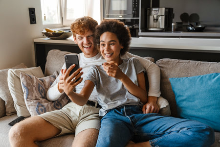 Young happy interracial couple taking selfie with phone together , while sitting on couch in cozy living room at homeの写真素材