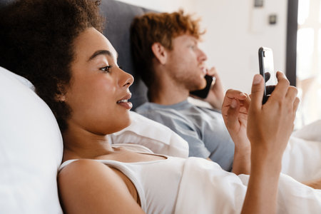 Young beautiful interracial couple holding and using their phones, while lying in bed together. Handsome redhead serious guy talking phone and his beautiful african curly girlfriend scrolling social mediaの写真素材