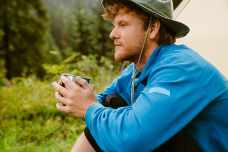 Young white tourist man drinking tea from metal cup while sitting at tent in forestの写真素材