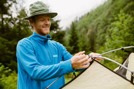 Smiling young ginger man wearing panama setting up tent during hiking in green forest