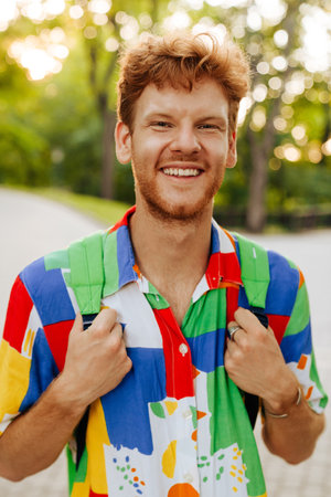 Young handsome smiling happy man in colorful shirt with backpack looking at camera, while standing in parkの写真素材