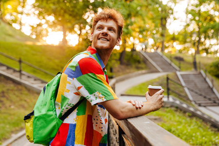 Young handsome smiling happy man in colorful shirt with coffee looking aside, while leaning on...