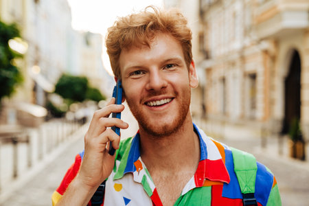 Portrait of young handsome smiling happy redhead man talking phone and looking at camera, while standing on the streetの写真素材