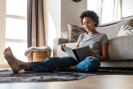 Young curly woman wearing jeans reading book while sitting on floor at homeの写真素材