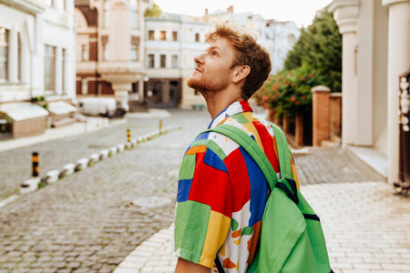 Portrait of young handsome calm redhead man in shirt with backpack looking aside, while standing with his back to camera on the streetの写真素材