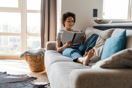 Young curly woman wearing jeans reading book while resting on sofa at homeの写真素材