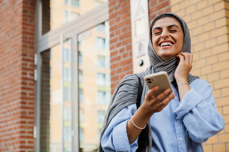 Cheerful middle-aged muslim woman wearing hijab using smartphone while standing outdoors at city streetの写真素材
