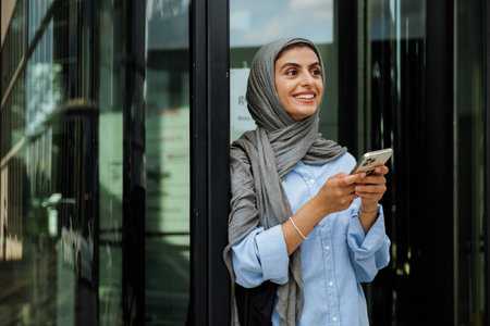 Cheerful middle-aged muslim woman wearing hijab using phone while standing outdoorsの写真素材