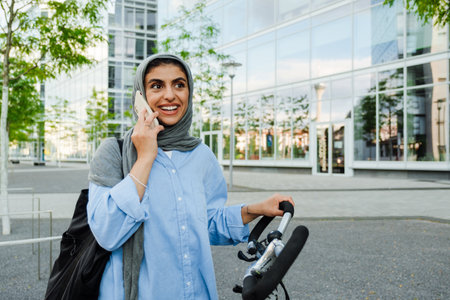 Young muslim businesswoman wearing headscarf using cellphone while standing by bicycle outdoorsの写真素材