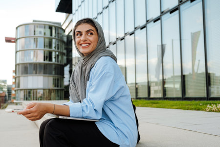Young muslim woman wearing headscarf using laptop and cellphone while sitting outdoorsの写真素材
