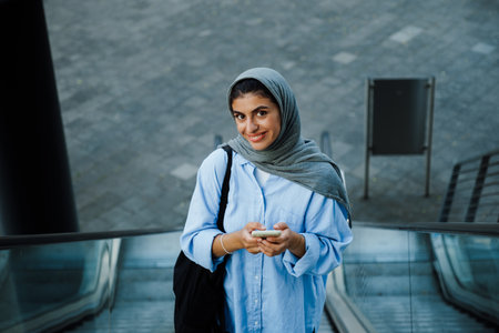 Young muslim woman wearing headscarf using cellphone while standing on escalator outdoorsの写真素材