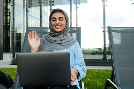 Young muslim woman wearing headscarf using laptop while sitting on chair outdoorsの写真素材