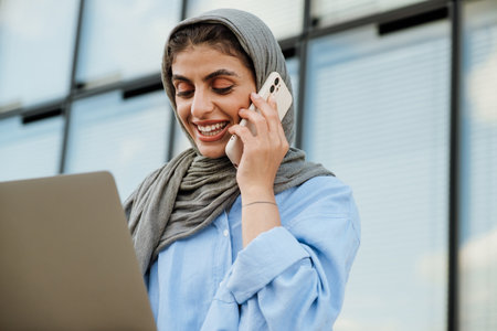 Cheerful middle-aged muslim woman wearing hijab talking on cellphone and using laptop while sitting outdoors at city streetの写真素材