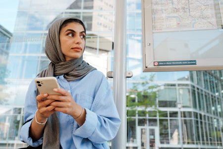 Young muslim woman wearing headscarf using cellphone while sitting on bus stop outdoorsの写真素材