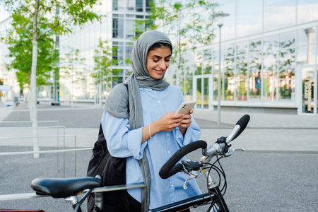 Young muslim businesswoman wearing headscarf using cellphone while standing by bicycle outdoorsの写真素材