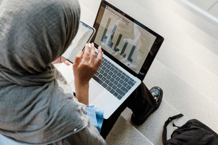 Young muslim woman wearing headscarf using cellphone and laptop while sitting on stairs outdoorsの写真素材