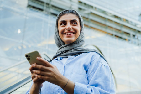 Young muslim woman wearing headscarf using cellphone while standing on escalator outdoorsの写真素材