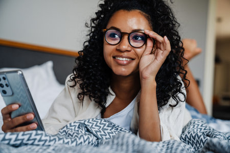 Black young woman in eyeglasses using mobile phone while resting on bed at homeの写真素材