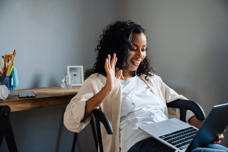 Black young woman waving hand and working with laptop while sitting on chair at homeの写真素材