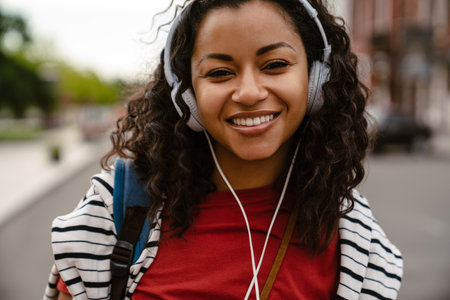 Young black woman in headphone looki camera while walking outdoorsの写真素材