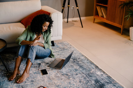 Cheerful young african woman having video chat using laptop while sitting on the floor at homeの写真素材