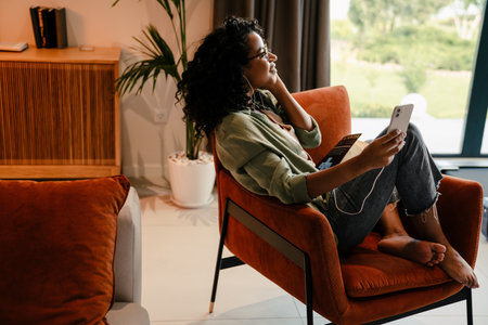 Profile of young smiling african woman in earphones using smartphone while sitting on armchair at homeの写真素材