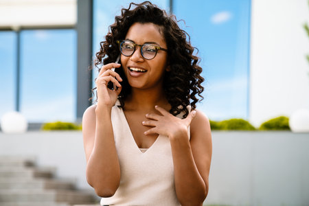 Cheerful young african woman in eyeglasses talking on mobile phone and gesturing while standing...