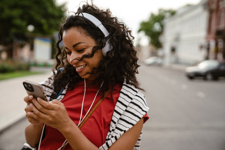 Young black woman in headphones using mobile phone while walking outdoorsの写真素材