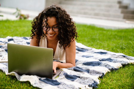 Young cheerful african woman in earphones using laptop while lying on blanket on green grassの写真素材