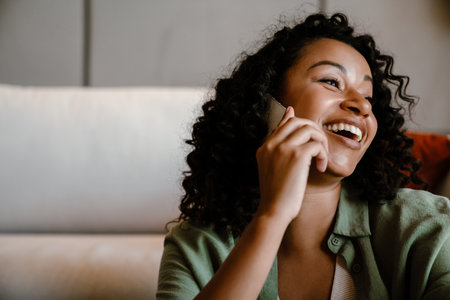 Excited young smiling african woman talking on mobile phone at homeの写真素材