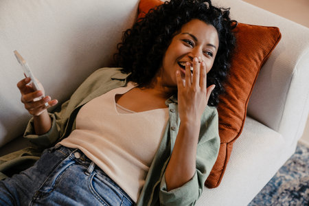 Cheerful young african woman using smartphone and gesturing while lying on sofa in cozy apartmentの写真素材