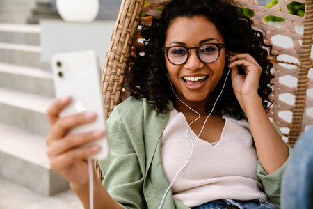 Excited smiling young african american woman in earphones using smartphone while sitting outdoors on rattan hang chairの写真素材