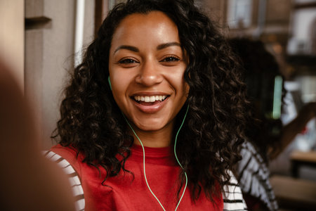 Young black woman in earphones taking selfie photo while sitting at cafe outdoorsの写真素材