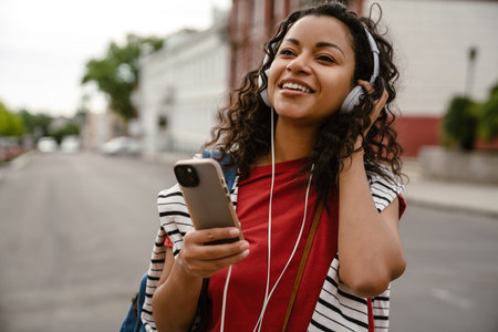 Young black woman in headphones using mobile phone while walking outdoorsの写真素材