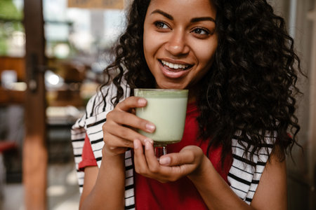 Young black woman smiling and drinking matcha latte while sitting at cafe outdoorsの写真素材