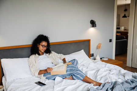 Black young woman in eyeglasses reading book while resting on bed at homeの写真素材
