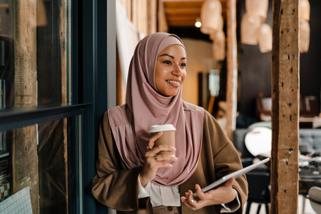 Cheerful arabian woman wearing hijab holding cup of coffee and tablet while standing in modern officeの写真素材