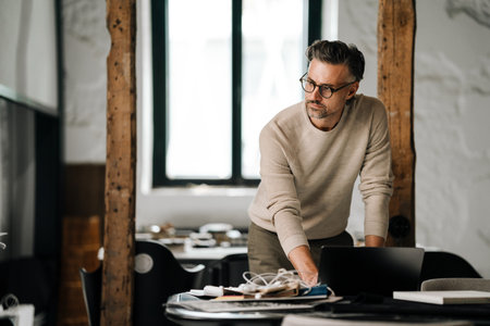 Pensive white middle-aged man in eyeglasses working with laptop in officeの写真素材
