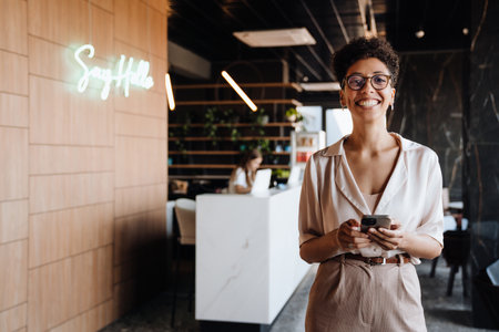 Happy black businesswoman smiling and using cellphone while working in office lobbyの写真素材