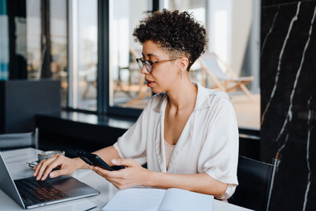 Middle-aged businesswoman using cellphone and working on laptop by table in officeの写真素材