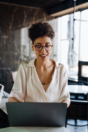 Happy black businesswoman smiling and using laptop while working in office lobbyの写真素材