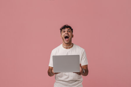 Young handsome enthusiastic man with opened mouth holding laptop and looking upward , while standing...