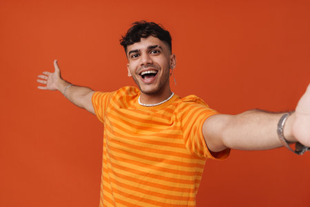Brunette young man wearing t-shirt gesturing while taking selfie photo isolated over red backgroundの写真素材