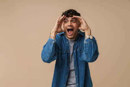 Young handsome stylish shocked man with opened mouth and wide opened eyes touching his head and looking aside, while standing over isolated brown backgroundの写真素材