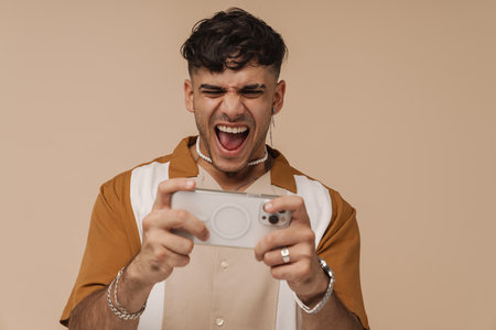 Young handsome stylish enthusiastic squinting man playing mobile phone with opened mouth, while standing over isolated brown backgroundの写真素材