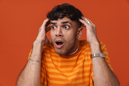 Brunette young man wearing t-shirt expressing surprise at camera isolated over red backgroundの写真素材
