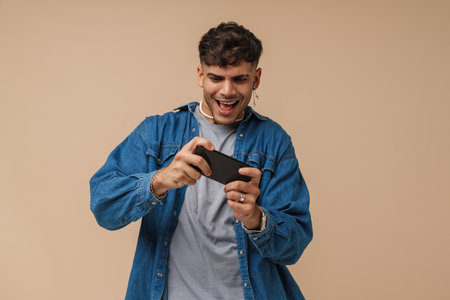 Young handsome stylish interested focused man with opened mouth playing mobile game on his phone, while standing over isolated brown backgroundの写真素材
