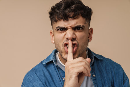 Brunette young man grimacing and showing silence gesture at camera isolated over beige backgroundの写真素材