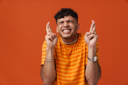 Brunette young man holding fingers crossed for good luck isolated over red background